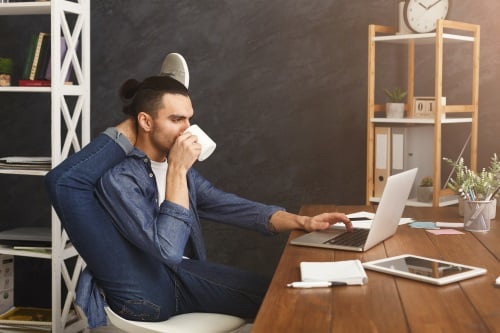 Flexibler Mann beim Arbeiten Mann mit Tasse in der Hand und dem rechten Bein hinter seinem Kopf vor dem Laptop